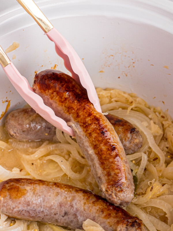 Tongs picking up a bratwurst from a crockpot with sauerkraut in it.