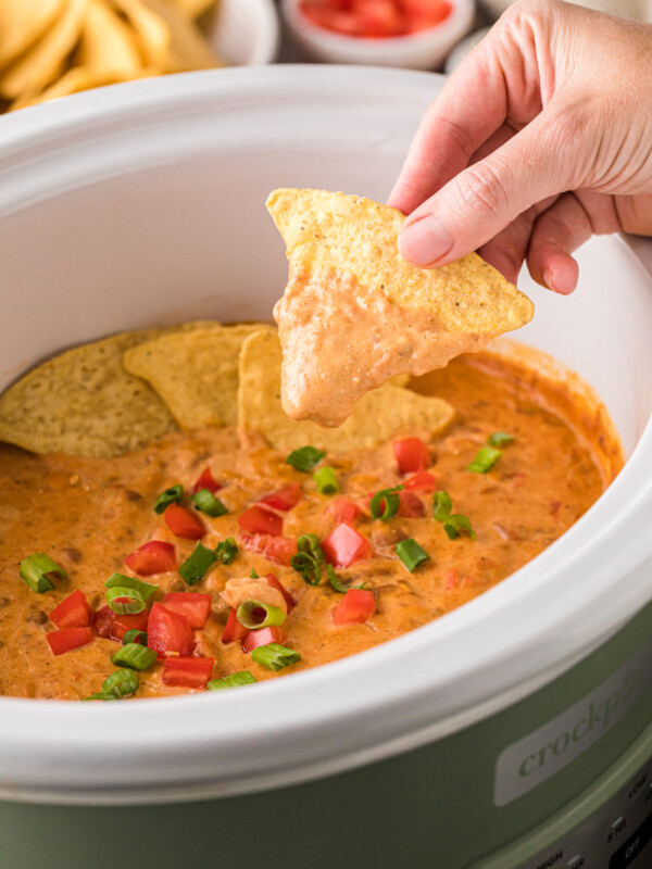 A hand holding a tortilla chip covered in dip over a crockpot.