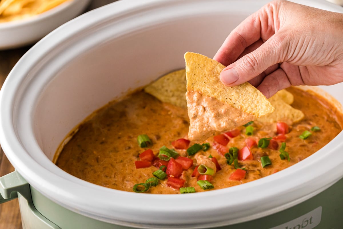 A hand holding a dip-covered tortilla chip over a crockpot of dip.