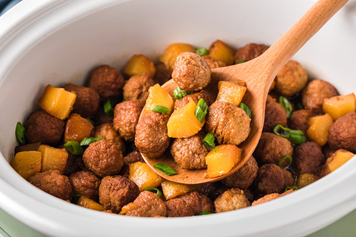 A wooden spoon holding a scoop of pineapple meatballs over a crockpot.